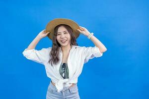 Porträt von jung asiatisch Frau im beiläufig Kleidung zum Ferien und Strand Stimmung berühren ihr Stroh Hut isoliert auf Blau Hintergrund zum Reise und Mode Studio Schnitt aus Konzept foto