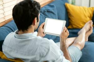 gut aussehend Mann ist mit ein Digital Tablette während ruhen auf Couch beim heim. Mann Sitzung auf Sofa und halten Tablette Computer mit isoliert Bildschirm foto