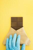 Bar von Milch Schokolade im ein Verpackung ist gehaltenen durch ein Hand im ein medizinisch Handschuh auf ein Gelb Hintergrund. Süße. Welt Schokolade Tag 2020 während das Coronavirus. foto