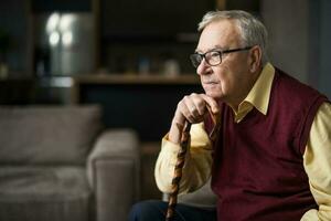 nachdenklich und besorgt Senior Mann ist Sitzung auf Bett im seine heim. Porträt von Senior Mann mit Gehen Stock. foto