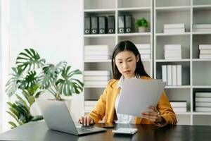 asiatisch Geschäftsfrau Arbeiten im das modern Büro mit Arbeiten Notizblock, Tablette und Laptop Unterlagen foto