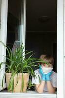 Quarantäne von ein Kind von COVID-19. ein amerikanisch Junge im medizinisch Handschuhe sieht aus aus das Fenster. foto