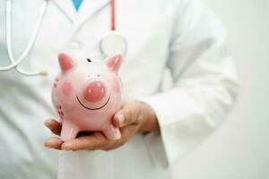 asiatisch Frau Arzt halten Schweinchen Bank, Kosten von Behandlung oder Bildung Konzept. foto