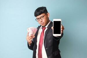 Porträt von jung asiatisch Geschäft Mann im beiläufig passen zeigen Telefon und halten tausend Rupien suchen beim Kamera mit ernst Gesicht. isoliert Bild auf Blau Hintergrund foto