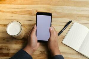 schließen oben von männlich Hand mit Handy, Mobiltelefon Telefon im leer Bildschirm mit leeren Notizbuch und ein Tasse von Kaffee während Sitzung beim Cafe foto