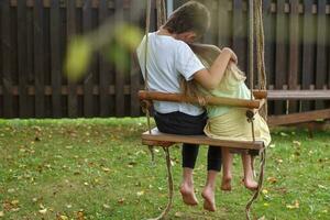 Kinder sitzen auf einer Schaukel im Garten. älterer bruder umarmt kleine schwester foto