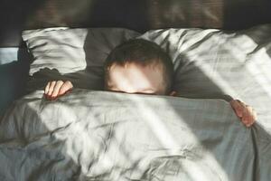 Junge schläft im Bett. Kind liegt auf Kissen und bedeckt sein Gesicht mit einer Decke foto