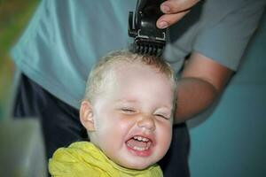 das zuerst Haarschnitt auf das Kopf von ein wenig Junge im seine Leben. ein Mann ist ein Vater Wer schneidet seine Söhne Haar. rasieren das Locken aus das Babys Kopf mit ein elektrisch Rasierer foto