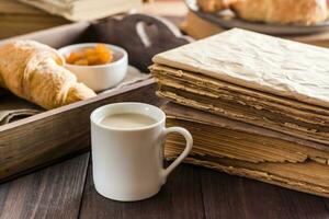 ein Tasse von Kaffee Nächster zu ein Stapel von alt Bücher und ein Tablett mit Croissants auf ein hölzern Tisch. Entspannung, Wellness und Zuhause Lektüre. foto