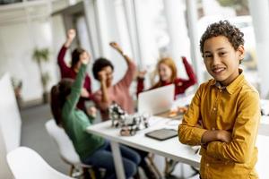 süßer kleiner Junge, der vor Kindern steht, die elektrisches Spielzeug und Roboter am Robotikklassenzimmer programmieren foto