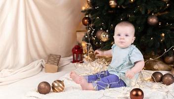 Ein Baby in Jeans sitzt in der Nähe eines Weihnachtsbaumes und spielt mit einer Girlande, in die er verwickelt ist foto