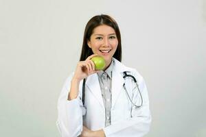 jung asiatisch weiblich Arzt tragen Schürze Uniform Tunika Stethoskop halten zeigen zeigen Essen gesund Grün Apfel foto