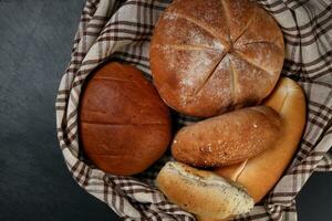 frisch gebacken Brot Laib Brötchen rollen runden lange mischen Wahrheit eingewickelt im kariert Küche Stoff Serviette Handtuch Über schwarz Hintergrund foto