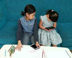 zwei wenig Süd Osten asiatisch Mädchen Kind malaiisch Chinesisch Studie abspielen zeichnen verwenden Clever Telefon auf Blau Stoff Sofa Sie sind autistisch und Nieder Syndrom foto