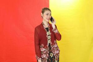 asiatisch Frau traditionell kastanienbraun Kebaya Sharong auf rot Gelb Papier Hintergrund sich unterhalten verwenden Telefon Smartphone foto