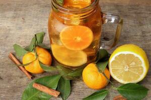Flüssigkeit Eis Zitrone Orange Tee mit Scheibe Grün Blatt Zimt Stock im transparent Glas Krug Becher auf rustikal Holz Hintergrund ganze Obst foto