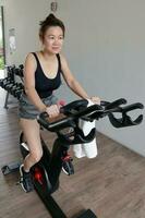 schön attraktiv Mitte Alter Süd-Ost asiatisch Frau ausüben stationär Fahrrad Zyklus Fahrrad im ein Gymnasium gesund Lebensstil aussehen nach vorne foto