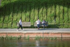 Vilnius, Litauen 03 06 2022 zwei Männer Sitzung auf ein Bank und ein Paar Gehen entlang das Fluss Bank im Sonnenuntergang Sonnenlicht foto