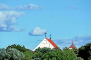 rot Dach oben von Christian Kirche mit Kreuz Über Grün Bäume auf Blau Himmel Hintergrund mit Weiß Wolken foto