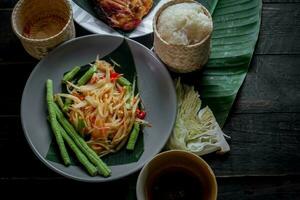 thailändisch Essen Gericht beide im Thailand und Asien, Papaya Salat oder wie wir Anruf es somtum ist ergänzt mit gegrillt Hähnchen und klebrig Reis mit frisch Pfannengerichte. serviert auf das schwarz hölzern Tisch. foto