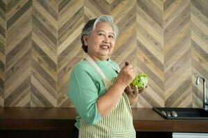 Porträt von glücklich Senior asiatisch Frau geduldig Essen frisch und sauber Vegetarier Salat im das Küche beim heim, Kochen gesund Essen beim heim, vorbereiten Mahlzeiten beim heim. Diät und aktiv Senioren Konzept foto