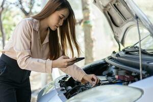 wütend asiatisch Frau mit ein Smartphone Video Konferenz zum Hilfe nach ein Auto Nervenzusammenbruch auf Straße. Konzept von ein Fahrzeug Motor Problem oder Unfall und Notfall Hilfe von ein Fachmann Mechaniker foto