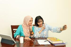 zwei jung asiatisch malaiisch Muslim Frau tragen Kopftuch beim Zuhause Büro Schüler Sitzung beim Tabelle Telefon Computer Buch dokumentieren Selfie selbst Porträt mit Smartphone foto