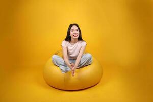 schön jung Süd Osten asiatisch Frau sitzt auf ein Gelb Sitzsack Sitz Orange Gelb Farbe Hintergrund Pose Mode Stil elegant Schönheit Stimmung Ausdruck sich ausruhen entspannen foto