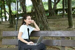 jung asiatisch Chinesisch Frau draussen auf Park Bank verwenden Laptop Computer denken lesen aussehen glücklich Wunder foto