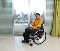 jung Mann Süd Osten asiatisch malaiisch Ursprung Sitzung auf Rollstuhl gegen Fenster Licht. gelähmt Taille Nieder fällig zu Unfall foto