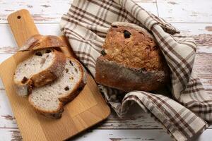 rustikal ganze Korn Handwerker Brot Laib Scheibe mit Cranberry Rosine trocken Obst Nüsse eingewickelt im Dame Stoff mit hölzern Hacken Tafel Über Tabelle oben eben legen Aussicht foto