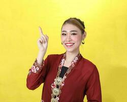 asiatisch Frau traditionell rot kastanienbraun Kebaya Sharong auf Gelb Papier Hintergrund posieren Finger zeigen oben foto