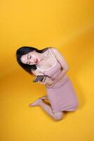 schön jung Süd Osten asiatisch Frau sitzen Nieder auf Fußboden Gelb Orange Farbe Hintergrund halt verwenden Text Clever Telefon Pose Mode Stil elegant Schönheit Stimmung foto