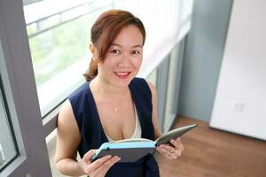 schön attraktiv Mitte Alter Süd-Ost asiatisch Frau Zuhause Büro Arbeit lesen sehen dokumentieren Buch Hinweis Stehen in der Nähe von Glas Fenster aussehen beim Kamera städtisch Tageslicht Fenster Hintergrund foto