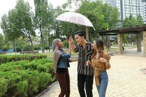 jung asiatisch malaiisch Mann Frau draussen Grün Park gehen sich unterhalten diskutieren mischen unter Regenschirm foto