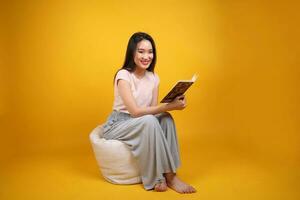 schön jung Süd Osten asiatisch Frau sitzt auf ein Weiß Sitzsack Sitz Orange Gelb Farbe Hintergrund Pose Mode Stil elegant Schönheit Stimmung Ausdruck entspannen Lächeln aussehen lesen Buch foto