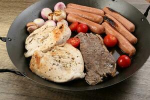 mischen Treffen schwenken Hähnchen Brust Rindfleisch Steak Würstchen Tomate Zwiebel Knoblauch Öl braten Grill auf Metall braten schwenken zwei Holz Griff auf rustikal hölzern Hintergrund foto