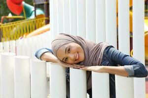 jung asiatisch malaiisch Muslim Frau Kopftuch draussen Grün Pose Kopf auf Hand ruhen auf Pole foto