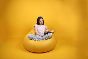 schön jung Süd Osten asiatisch Frau sitzt auf ein Gelb Sitzsack Sitz Orange Gelb Farbe Hintergrund Pose Mode Stil elegant Schönheit Stimmung Ausdruck sich ausruhen entspannen lesen Buch denken foto
