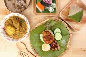 nasi Lemak duftend Reis Kokosnuss Milch mit Sambal Freund Erdnuss Anschovis Ei verpackt Banane Blatt runden Bambus Teller bunt Nyonya kuih Süss Wüste Palme Blatt Teller auf hölzern Hintergrund foto