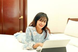 jung asiatisch malaiisch Muslim Frau tragen Baju Kurung Kleid beim Zuhause auf Bett Studie Arbeit auf Computer Tab aussehen beim Kamera foto