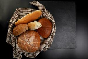 frisch gebacken Brot Laib Brötchen rollen runden lange mischen Wahrheit eingewickelt im kariert Küche Stoff Serviette Handtuch Über schwarz Hintergrund foto