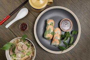 Vietnamesisch Garnele Garnele sehen durch Reis Papier Frühling rollen poh Rindfleisch Nudeln Suppe Rindfleisch Ball Bruststück Gekröse Bauch Teile thailändisch Basilikum Limette rustikal hölzern Tabelle Essstäbchen Löffel Soße dunkel Teller Zinn Becher foto