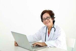 Alten asiatisch Chinesisch weiblich Arzt auf Weiß Hintergrund tragen Weiß Schürze Stethoskop aussehen beim Kamera Tippen Arbeiten beim Laptop Notizbuch pc foto