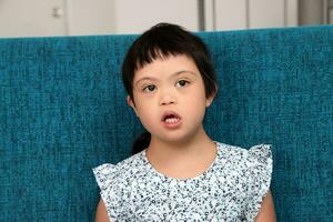 Süd-Ost asiatisch klein wenig Mädchen Kind Sitzung auf Blau Sofa aussehen glücklich Lächeln Pose. sie haben Nieder Syndrom foto