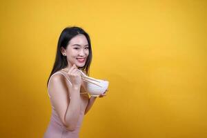 schön jung Süd Osten asiatisch Frau halten chines leeren Stäbchen Chinesisch Suppe Löffel Schüssel Utensil so tun als ob Schauspielkunst posieren sehen Essen Geschmack Geruch Futter Angebot Befriedigung lecker Gelb Orange Hintergrund foto