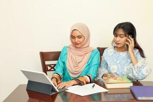 zwei jung asiatisch malaiisch Muslim Frau tragen Kopftuch beim Zuhause Büro Schüler Sitzung beim Tabelle Telefon Computer Buch dokumentieren Studie sich unterhalten Arbeit mischen foto