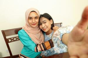 zwei jung asiatisch malaiisch Muslim Frau tragen Kopftuch beim Zuhause Büro Schüler Sitzung beim Tabelle Telefon Computer Buch dokumentieren Selfie selbst Porträt Umarmung Pflege Zuneigung pov Punkt von Aussicht foto