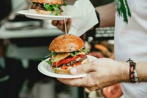 Mann hält im seine Hand Burger beim ein Burger Fest. Abendessen Hamburger Essen Fest Party. groß Burger im Hände beim Straße Essen draussen Festival. Festival Essen Tag foto