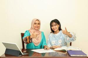 zwei jung asiatisch malaiisch Muslim Frau tragen Kopftuch beim Zuhause Büro Schüler Sitzung beim Tabelle sich unterhalten mischen Computer Buch dokumentieren Studie Daumen oben Zeichen aussehen beim Kamera foto